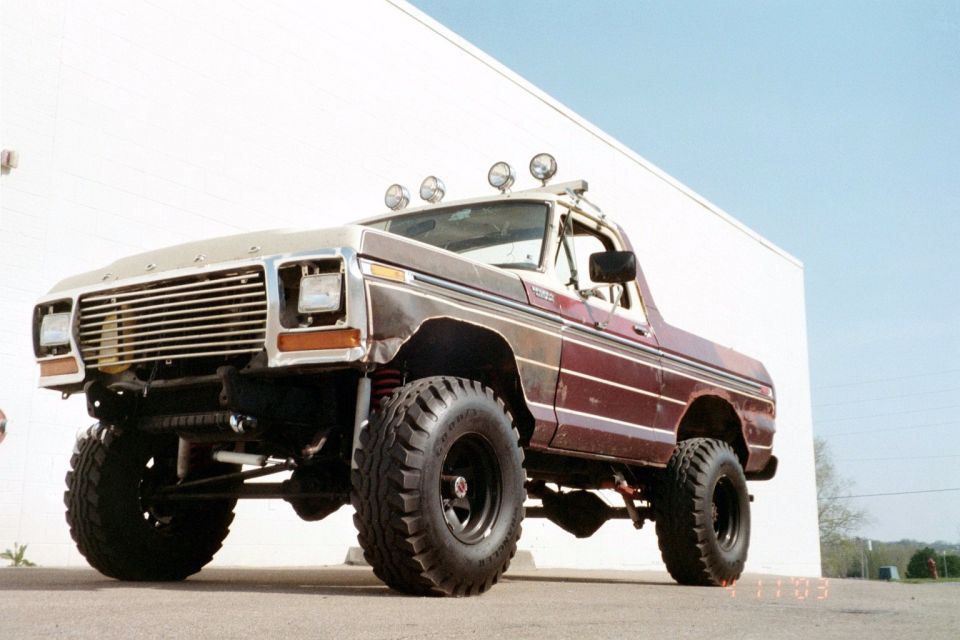 Lifted burgundy and tan Ford 4x4 pickup with roll bar and driving lights beside a white wall