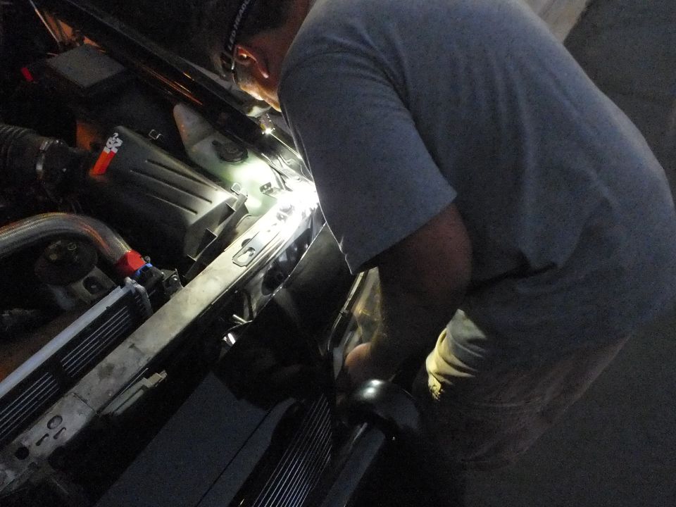 Worker leaning over radiator support and core of a vehicle illuminated at night