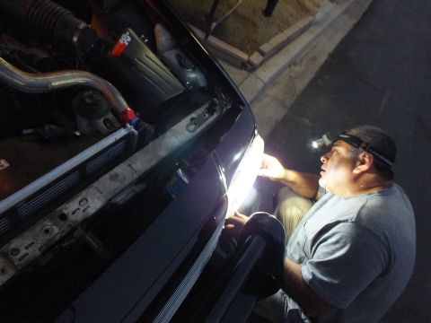 2003 Ford Crown Victoria - Bearded worker with a head lamp examines the front headlight corner of a dark car after dark
