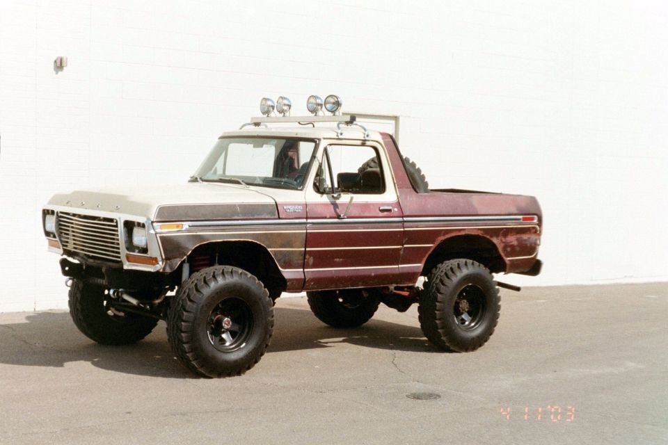 Side profile of a lifted burgundy and tan Ford 4x4 short-bed pickup with roll bar and spare tire