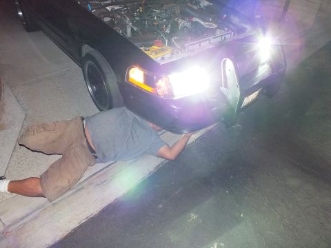 2003 Ford Crown Victoria - Worker lies on the pavement reaching beneath the front bumper of a dark car with headlights lit
