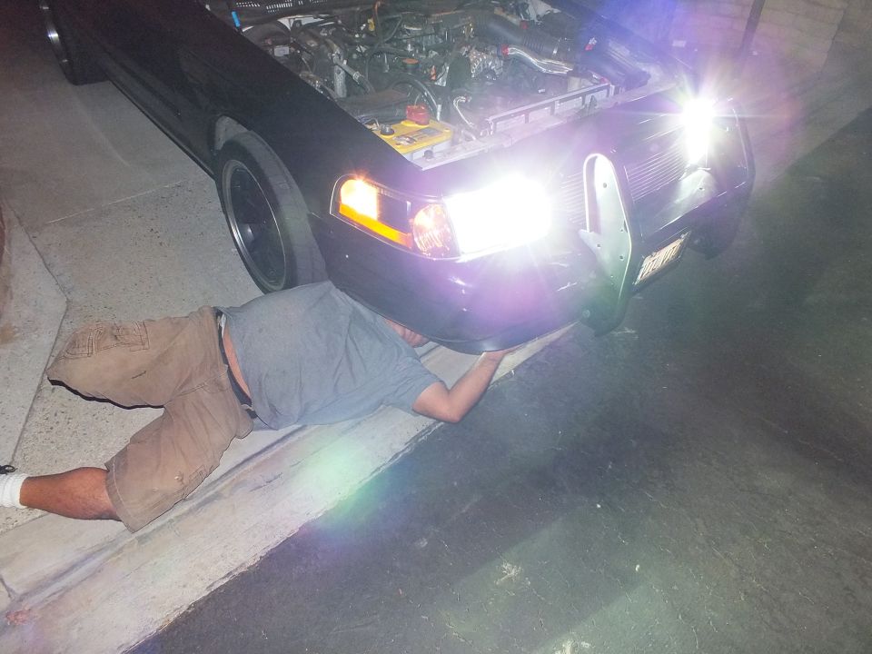 Man lying on ground reaching under front bumper of dark sedan with lights on