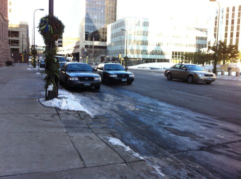 2003 Ford Crown Victoria - Two dark sedans are parked on a city street with snow and ice on the ground, while a gold sedan drives past.