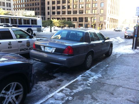 2003 Ford Crown Victoria - A 2003 Ford Crown Victoria is parked on a city street with snow and ice on the ground, next to a police car.