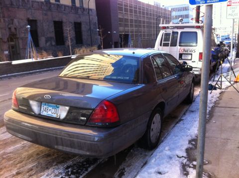 2003 Ford Crown Victoria - A dark colored Ford Crown Victoria police car is parked on a snowy street beside a news van.