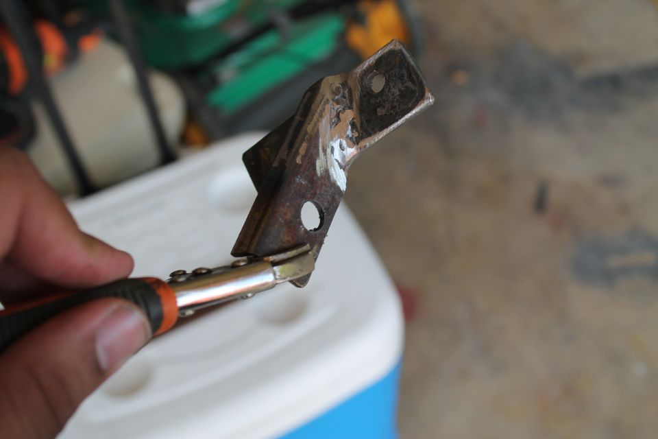 Hand holding pliers on a rusty metal bracket. Background shows a white cooler and garage floor