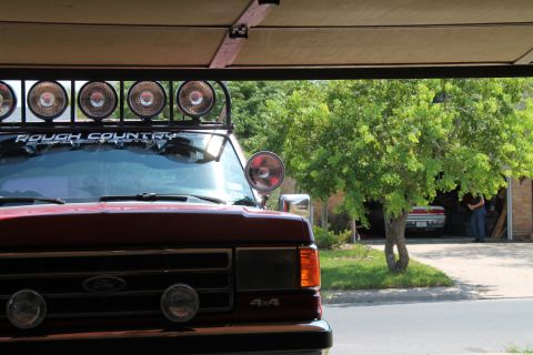 1990 Ford Bronco - A red Ford Bronco is parked under a cover, equipped with a light bar and auxiliary lights on the bumper.