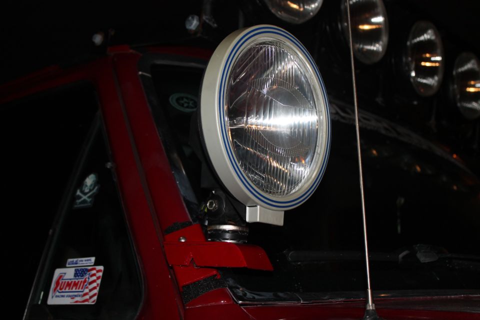 Close-up of an aftermarket auxiliary light with a blue stripe on its bezel, mounted on a red vehicle. A 'Summit' sticker is visible on the window