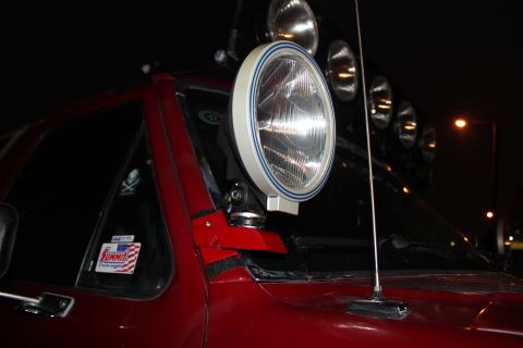 1990 Ford Bronco - A large auxiliary light is mounted on the A-pillar of a red vehicle, with additional lights on the roof rack.