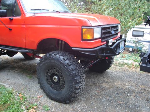 1989 Ford Bronco - Front three-quarter view of a red Ford Bronco lifted on large mud-terrain tires with black beadlock-style wheels and a black tubular front…