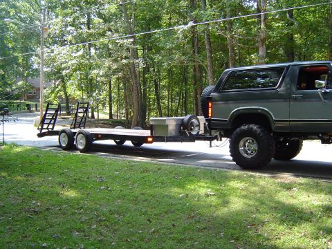 1986 Ford Bronco - Ford Bronco towing a flatbed trailer on a paved road with trees in the background.