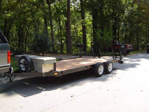 1986 Ford Bronco - A flatbed trailer is parked on a paved surface, with trees lining the background.