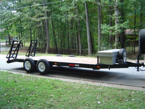 1986 Ford Bronco - A flatbed trailer is parked on the side of a road, ramps, a toolbox, and a spare tire mounted at the front.