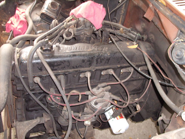 Dusty inline-six engine in a truck engine bay with black valve cover, pink rag on carburetor, and new white oil filter