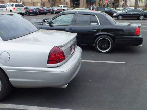 2003 Ford Crown Victoria - Silver and black full-size sedans on black aftermarket wheels parked back to back in a lot