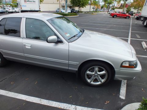 2003 Ford Crown Victoria - Profile of a silver four-door sedan on polished five-spoke wheels; storefronts and palms behind.