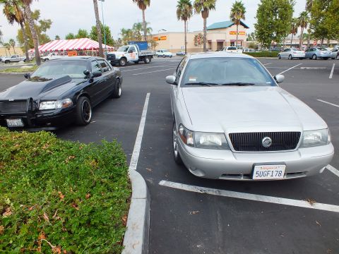 2003 Ford Crown Victoria - Front three-quarter of a silver Mercury sedan, plate 5UGF178, parked next to a black sedan with brush guard.