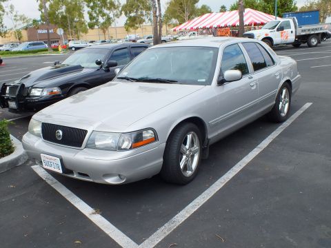 2003 Ford Crown Victoria - Front quarter of a silver Mercury sedan on polished wheels; a black sedan and red tent are behind.
