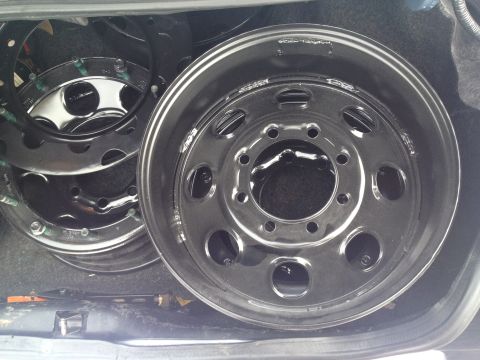 1989 Ford Bronco - Several shiny metal rims are stacked in the back of a vehicle, with a jack visible below.