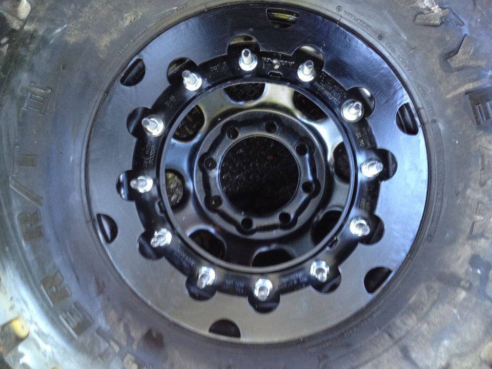 Close-up of a black off-road wheel with multiple lug nuts and a large tire. Supermotors.net logo in the corner