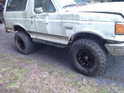 1989 Ford Bronco - A weathered white Ford Bronco is viewed from the front quarter, revealing its off-road tires and worn exterior.