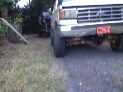 1989 Ford Bronco - A silver truck, possibly a Ford, is seen from the front, towing another vehicle on a trailer. A red license plate reads '1982'.