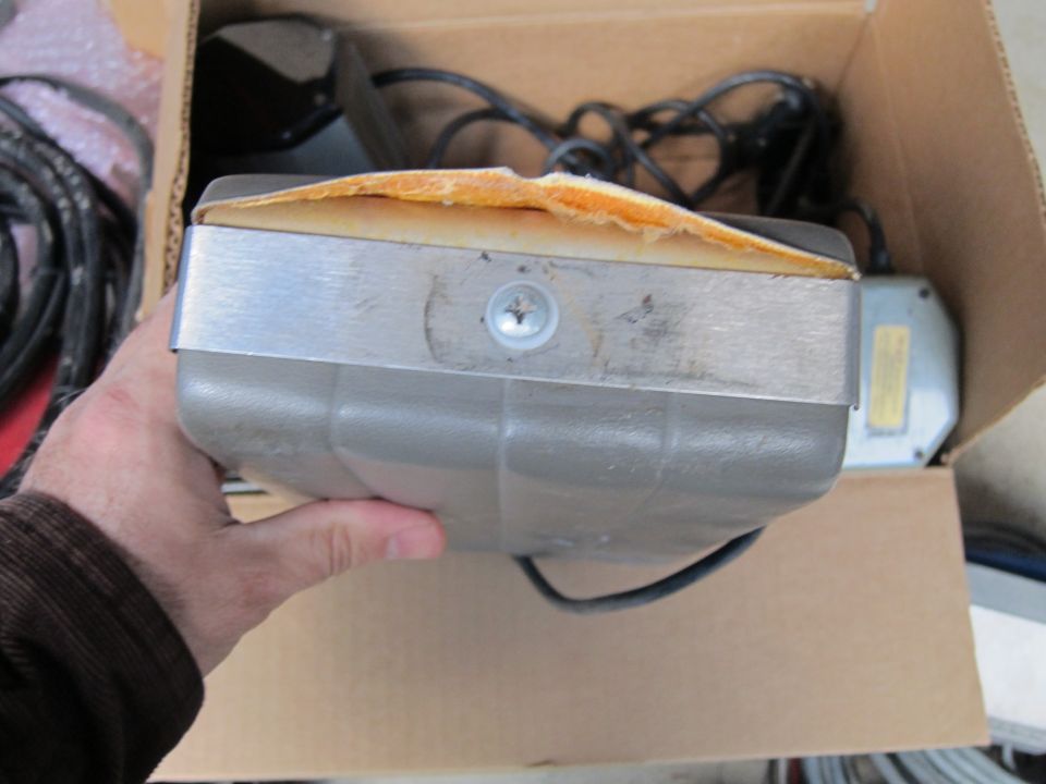 Gray electronic part with metal band and orange foam padding, held in a cardboard box