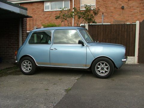 1980 MINI Cooper - A light blue 1980 MINI Cooper is parked in front of a brick house and wooden fence.