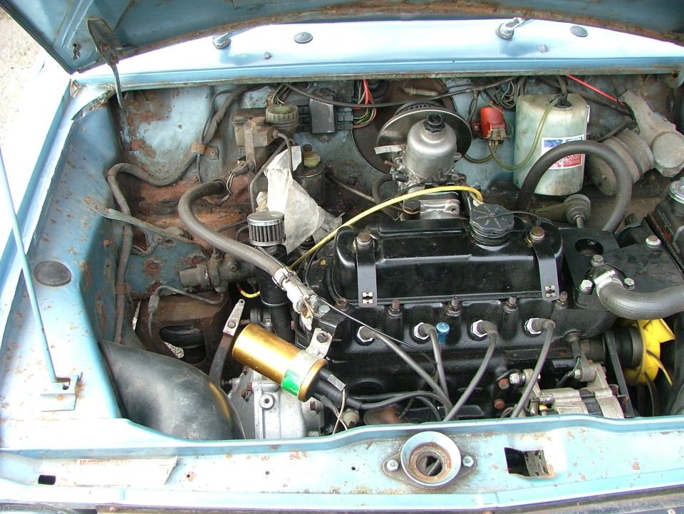 Engine bay of a light blue classic car, showing engine, hoses, and some rust on the inner fender