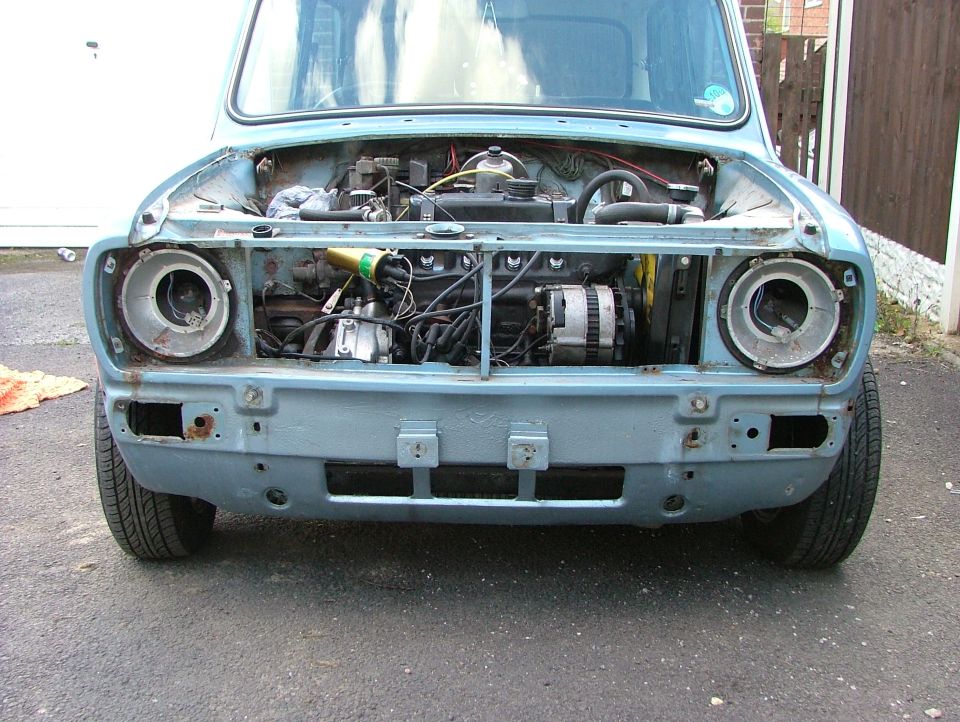 Light blue classic Mini with front panel off exposing engine, empty headlight buckets, surface rust.