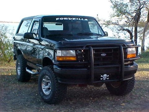 1995 Ford Bronco - A black Ford Bronco is parked on a dirt lot with trees and water in the background.