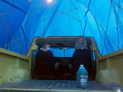 1978 Ford Bronco - Two children are seated in the back of a yellow pickup truck, which is covered by a large blue tarp.