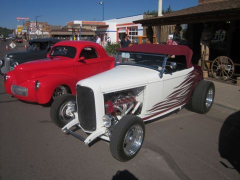 1991 Ford LTD Crown Victoria - A white and maroon hot rod with a visible engine and flame graphics is parked next to a red coupe at a car show.