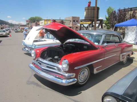1991 Ford LTD Crown Victoria - A red coupe with its hood open is displayed at an outdoor car show, with other vehicles and buildings in the background.