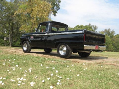 1966 Ford F-100 - A black pickup truck is parked outdoors on a dirt road with grass and trees. The truck has chrome accents and wheels.