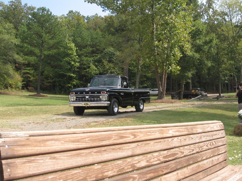 Black 1960s Ford F-100 pickup parked on gravel at a wooded rural property