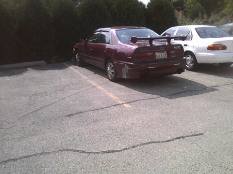 2003 Ford Crown Victoria - Maroon sedan with a large rear spoiler and aftermarket bumper damage is parked in a lot.