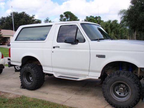 1995 Ford Bronco - A white Ford Bronco is parked on a concrete driveway, showing off its lifted suspension and large tires.