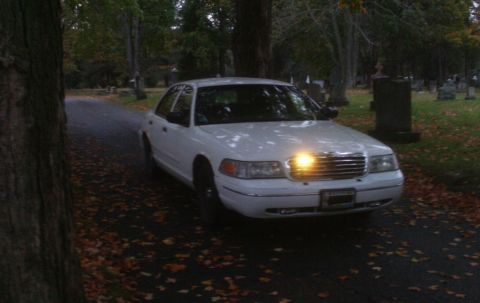 1998 Ford Crown Victoria - A white 1998 Ford Crown Victoria is parked on a road lined with trees and gravestones.