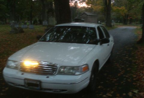 1998 Ford Crown Victoria - White 1998 Ford Crown Victoria parked on a road with fallen leaves, surrounded by trees and gravestones.