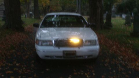 1998 Ford Crown Victoria - A white 1998 Ford Crown Victoria is parked in a graveyard, its grille emblem glowing.