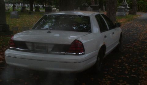 1998 Ford Crown Victoria - A white 1998 Ford Crown Victoria is parked on a road in a cemetery with gravestones visible in the background.