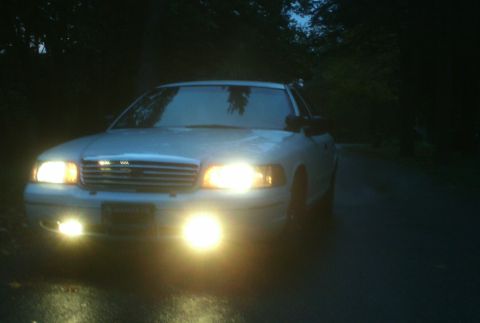 1998 Ford Crown Victoria - A white 1998 Ford Crown Victoria is seen from the front at night with its headlights illuminated.