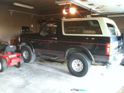 1994 Ford Bronco - A black Ford Bronco with a tan top is parked in a garage, with a red riding mower beside it.