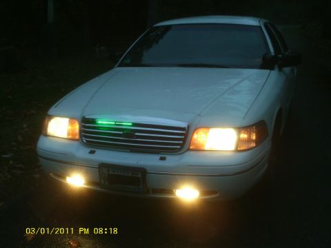 1998 Ford Crown Victoria - A white 1998 Ford Crown Victoria is shown from the front at night, with its lights on and a green light bar across the grille.