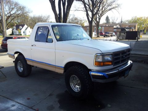 1996 Ford Bronco - A white pickup truck is parked outside on a paved surface, with trees and buildings in the background.