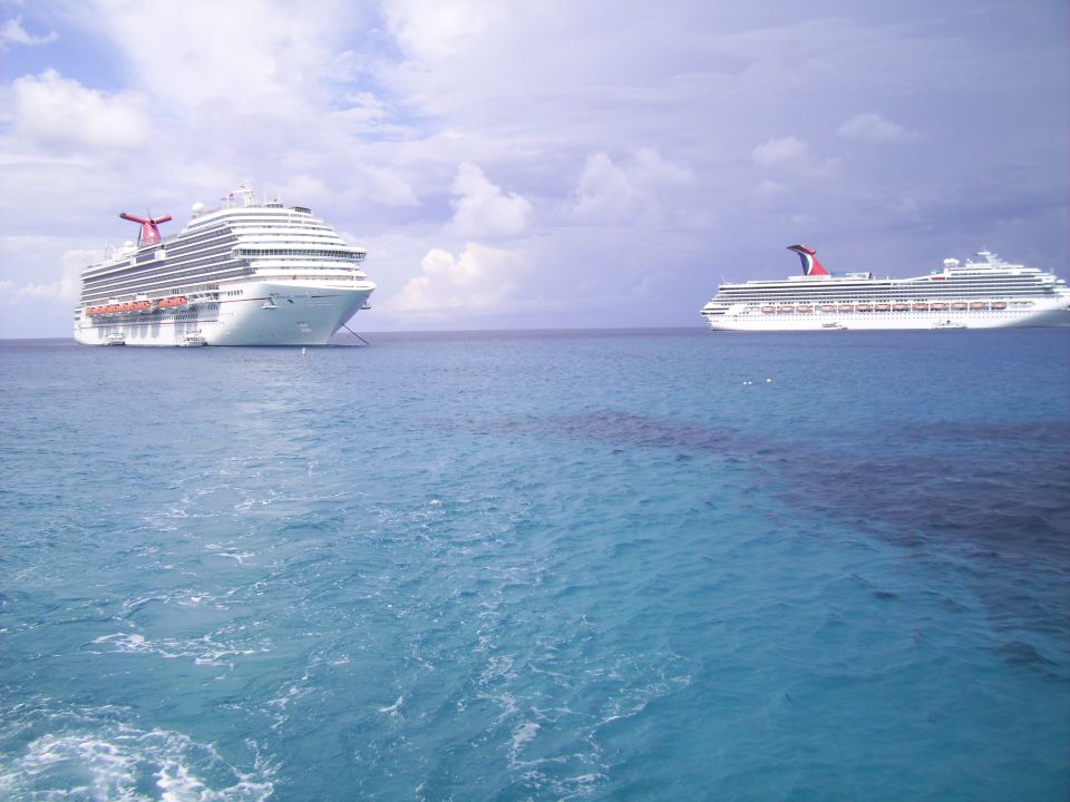 Two large white cruise ships float on a calm blue ocean under a cloudy sky