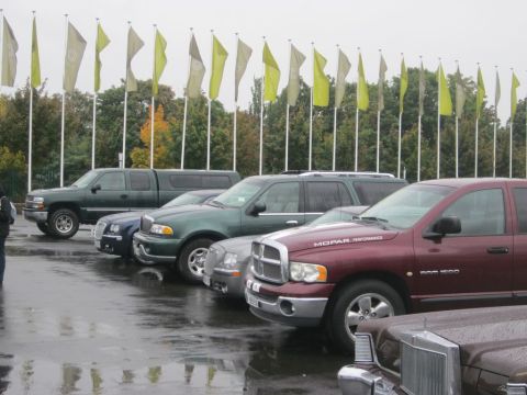 1991 Ford LTD Crown Victoria - Several vehicles are parked in a row under a line of yellow and tan flags, with trees in the background.