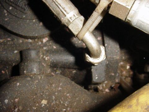 1992 Ford Bronco - Close-up of a metal fitting or fuel/AC line connection where a curved tube joins a hex-nut fitting on a grimy engine component. The surroun…