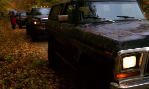 1979 Ford Bronco - A convoy of SUVs and trucks drives down a dirt road in autumn, with the lead vehicle being a dark Bronco.
